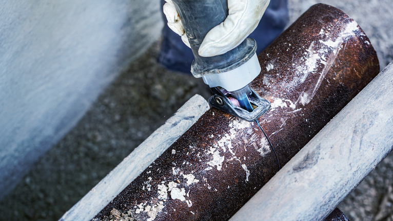 Seseorang yang mengenakan peralatan keselamatan menggunakan gergaji bolak-balik untuk memotong pipa logam berkarat.