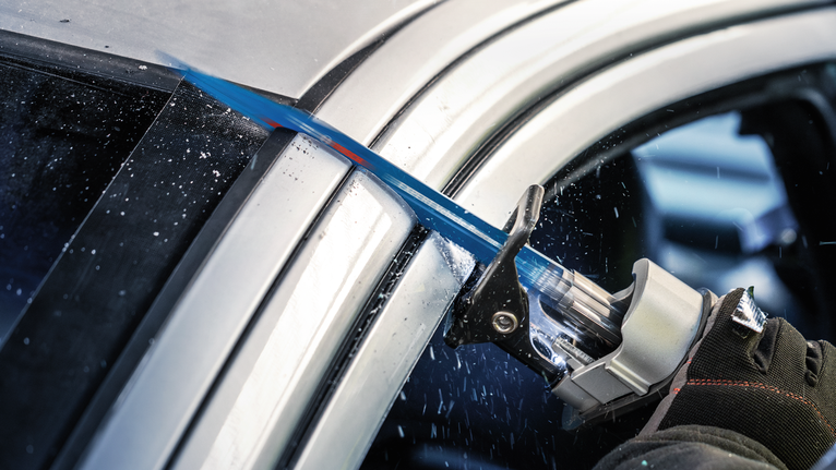 Seseorang yang mengenakan perlengkapan keselamatan menggunakan gergaji bolak-balik untuk memotong rangka mobil.