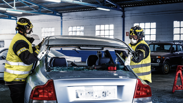 Dua pekerja yang mengenakan peralatan keselamatan melepas kaca depan dari sebuah mobil di dalam garasi.