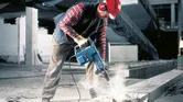 Seseorang yang mengenakan peralatan keselamatan mengoperasikan palu pembongkaran untuk memecah beton.
