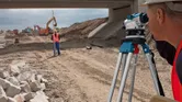 Orang yang mengenakan peralatan keselamatan menggunakan tripod tingkat survei di lokasi konstruksi.