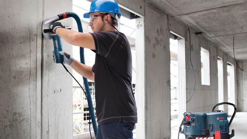 Seseorang yang mengenakan peralatan keselamatan sedang menggerinda dinding beton dengan alat penyedot debu di dekatnya.