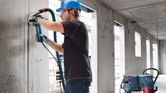 Seseorang yang mengenakan peralatan keselamatan sedang menggerinda dinding beton dengan alat penyedot debu di dekatnya.