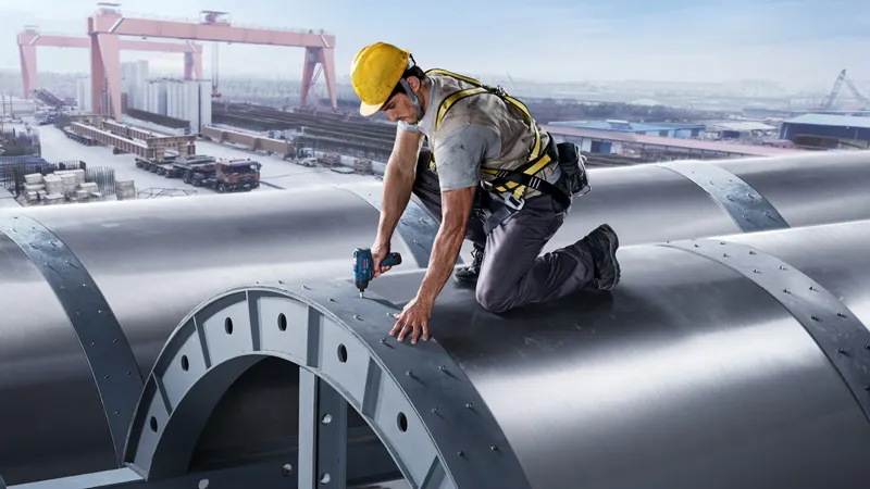 Seseorang yang mengenakan perlengkapan keselamatan mengencangkan baut pada struktur logam besar menggunakan perkakas listrik.