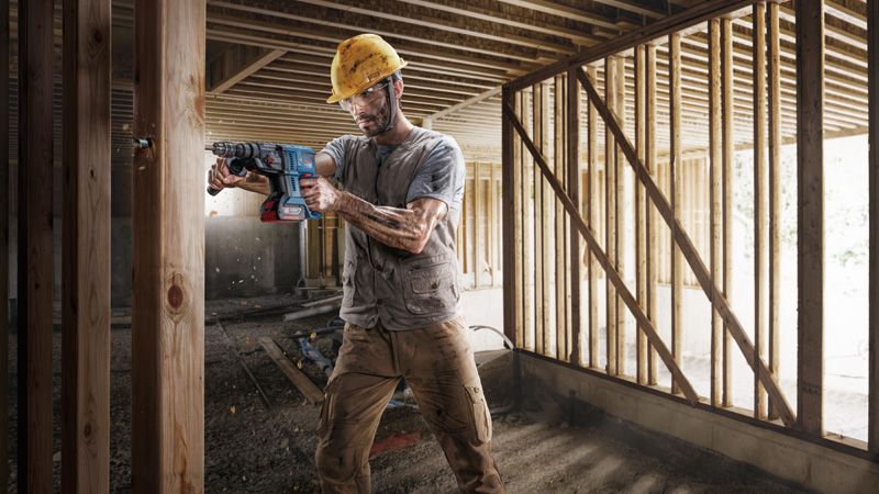 Seseorang yang mengenakan peralatan keselamatan mengebor balok kayu di lokasi konstruksi.