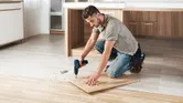 Seseorang mengebor papan kayu di lantai yang dikelilingi serutan kayu.