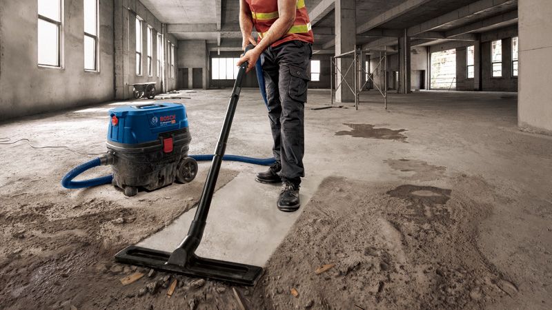 Seseorang yang mengenakan peralatan keselamatan sedang menyedot debu beton di sebuah bangunan besar yang belum selesai.