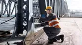 Seseorang yang mengenakan peralatan keselamatan mengebor balok baja di jembatan.