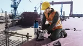 Seseorang yang mengenakan peralatan keselamatan mengoperasikan bor magnetik di platform galangan kapal.