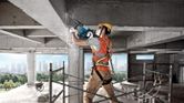 Seseorang yang mengenakan peralatan keselamatan menghancurkan tiang beton menggunakan palu pembongkaran.