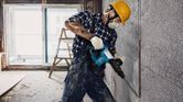 Seseorang yang mengenakan perlengkapan keselamatan menggunakan palu pembongkaran untuk menghancurkan tembok beton.