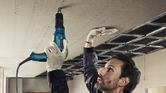 Seseorang yang mengenakan perlengkapan keselamatan mengencangkan drywall ke langit-langit menggunakan obeng.