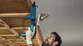 Seseorang yang mengenakan peralatan keselamatan memasang sekrup ke langit-langit dengan obeng drywall.