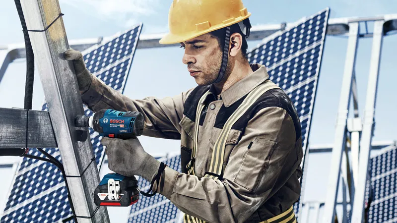 Seseorang yang mengenakan peralatan keselamatan menggunakan kunci pas benturan tanpa kabel pada rangka panel surya logam.