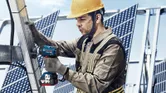 Seseorang yang mengenakan peralatan keselamatan menggunakan kunci pas benturan tanpa kabel pada rangka panel surya logam.