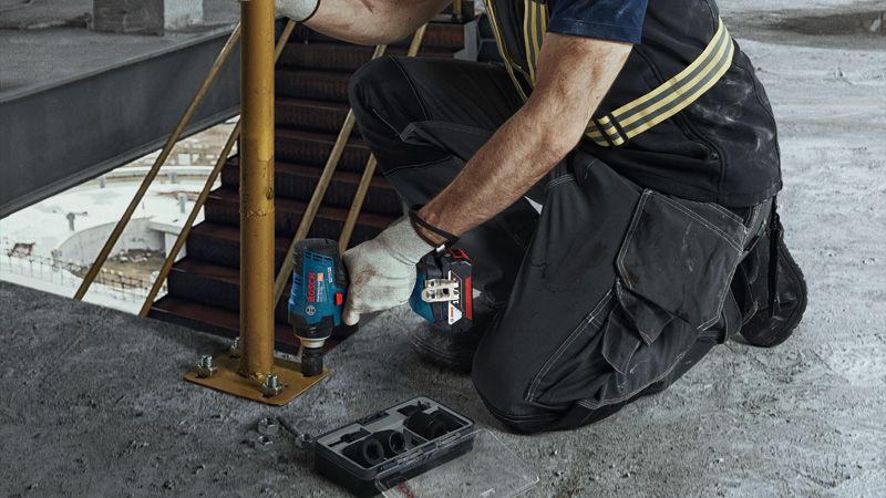 Seseorang yang mengenakan perlengkapan keselamatan mengencangkan baut pada tiang logam menggunakan kunci pas benturan tanpa kabel.