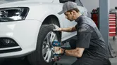 Seseorang yang mengenakan perlengkapan keselamatan menggunakan kunci pas benturan tanpa kabel untuk mengencangkan baut roda pada mobil.