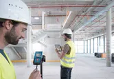 Dua orang yang mengenakan peralatan keselamatan menggunakan pengukur laser dan tablet di dalam gedung yang sedang dibangun.