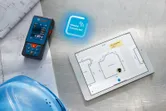 Pengukur laser di samping tablet digital yang menampilkan denah lantai di samping helm keselamatan biru.