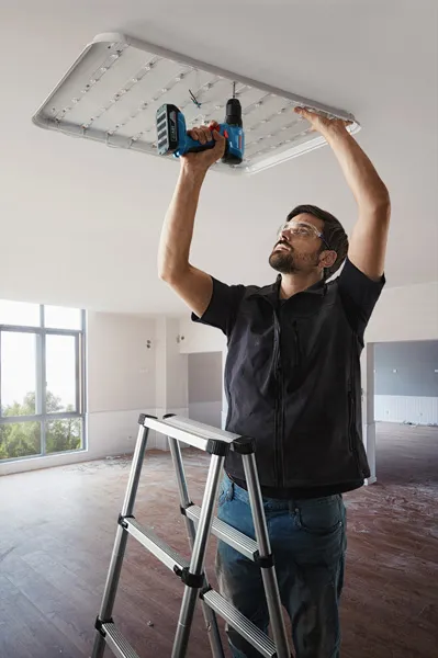 Seseorang yang mengenakan peralatan keselamatan memasang lampu langit-langit menggunakan bor listrik.