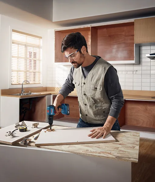 Orang yang mengenakan peralatan keselamatan mengebor papan kayu di ruang kerja dapur.