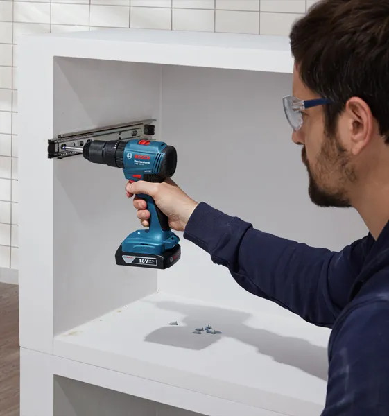 Seseorang yang mengenakan peralatan keselamatan menggunakan bor tanpa kabel untuk memasang rel kabinet.