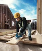 Seseorang yang mengenakan peralatan keselamatan memotong papan kayu dengan gergaji ukir nirkabel di lokasi konstruksi.