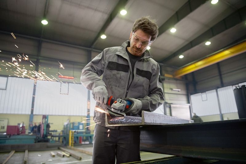 Seseorang yang mengenakan peralatan keselamatan menggiling balok baja, sehingga menghasilkan percikan api yang beterbangan.