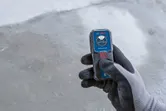 Orang yang mengenakan peralatan keselamatan memegang pengukur laser di lantai beton.
