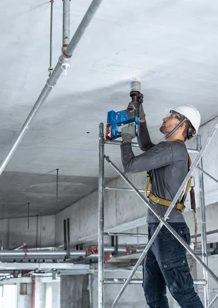 Seseorang yang mengenakan peralatan keselamatan mengebor langit-langit beton sambil berdiri di atas perancah.