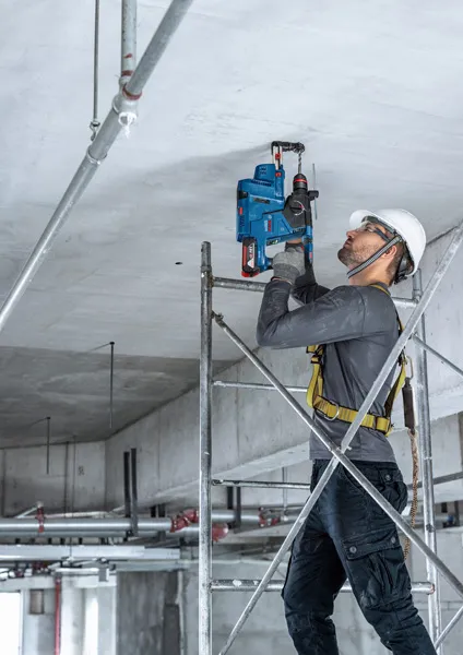 Seseorang yang mengenakan peralatan keselamatan mengebor langit-langit beton sambil berdiri di atas perancah.