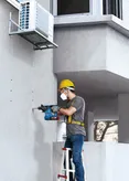 Seseorang yang mengenakan peralatan keselamatan mengebor lubang ke dinding beton di bawah AC.