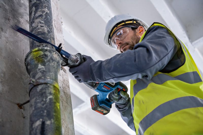 Seseorang yang mengenakan peralatan keselamatan memotong pipa logam dengan gergaji bolak-balik.