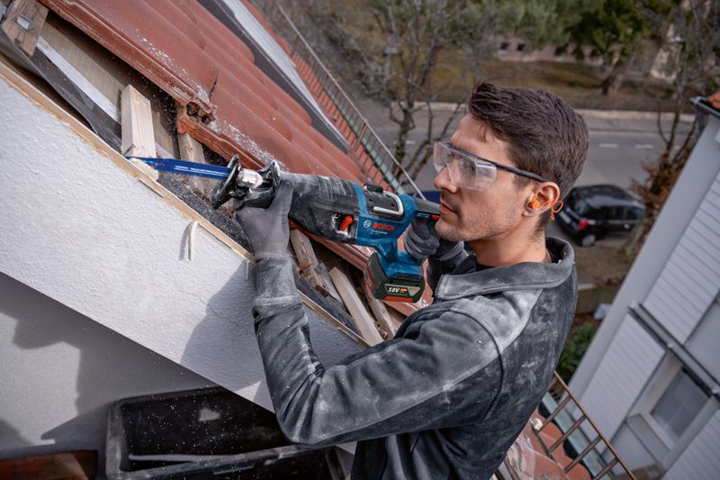 Seseorang yang mengenakan peralatan keselamatan menggunakan gergaji bolak-balik untuk memotong kayu di atap.