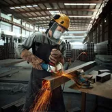 Seseorang yang mengenakan peralatan keselamatan menggiling balok logam dengan penggiling sudut, percikan api beterbangan.