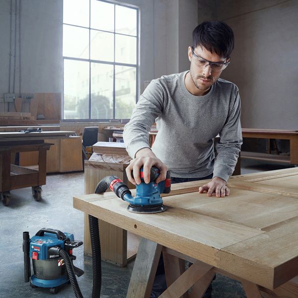 Seseorang yang mengenakan perlengkapan keselamatan mengampelas pintu kayu dengan sander orbital yang terhubung.