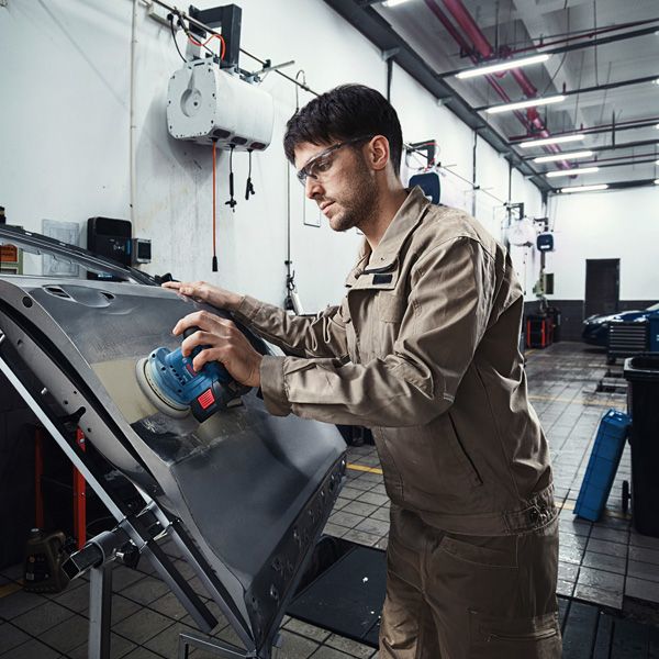 Seseorang yang mengenakan perlengkapan keselamatan mengampelas pintu mobil dengan mesin pengamplas.