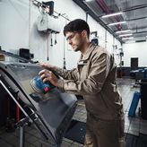 Seseorang yang mengenakan perlengkapan keselamatan mengampelas pintu mobil dengan mesin pengamplas.