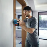 Seseorang yang mengenakan perlengkapan keselamatan sedang mengampelas dinding dengan mesin pengamplas listrik tanpa kabel.