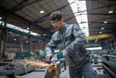 Seseorang yang mengenakan peralatan keselamatan menggiling logam, menciptakan percikan api di bengkel industri.