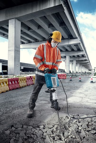 Seseorang yang mengenakan peralatan keselamatan memecahkan beton dengan pemutus arus listrik besar di luar ruangan.