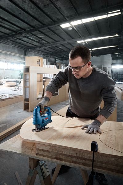 Orang yang mengenakan perlengkapan keselamatan menggunakan gergaji ukir untuk memotong bentuk lengkung pada kayu lapis.