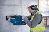 Seseorang yang mengenakan peralatan keselamatan mengebor dinding beton dengan palu putar tanpa kabel.