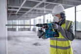 Seseorang yang mengenakan peralatan keselamatan mengebor beton menggunakan palu putar tanpa kabel.