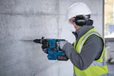 Seseorang yang mengenakan peralatan keselamatan mengebor dinding beton dengan palu putar tanpa kabel.