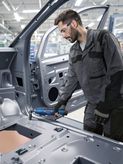 Orang yang mengenakan perlengkapan keselamatan menggunakan perkakas listrik pada rangka mobil di lingkungan industri.