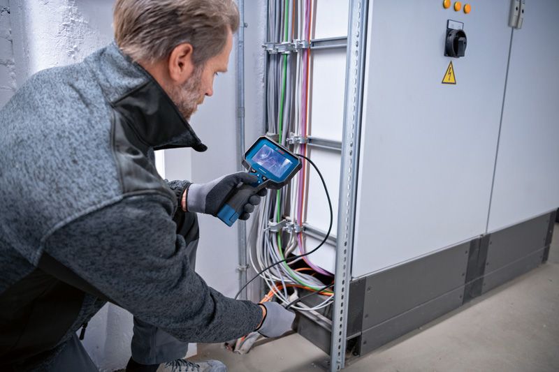 Seseorang yang mengenakan peralatan keselamatan memeriksa kabel listrik menggunakan kamera inspeksi.