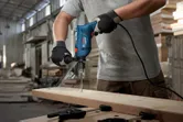 Seseorang yang mengenakan perlengkapan keselamatan mengebor papan kayu dengan bor tumbukan.
