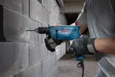 Seseorang yang mengenakan peralatan keselamatan mengebor dinding blok beton dengan bor tumbukan.