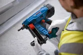 Seseorang yang mengenakan perlengkapan keselamatan sedang memasang paku ke dalam alat pemaku beton tanpa kabel berwarna biru.
